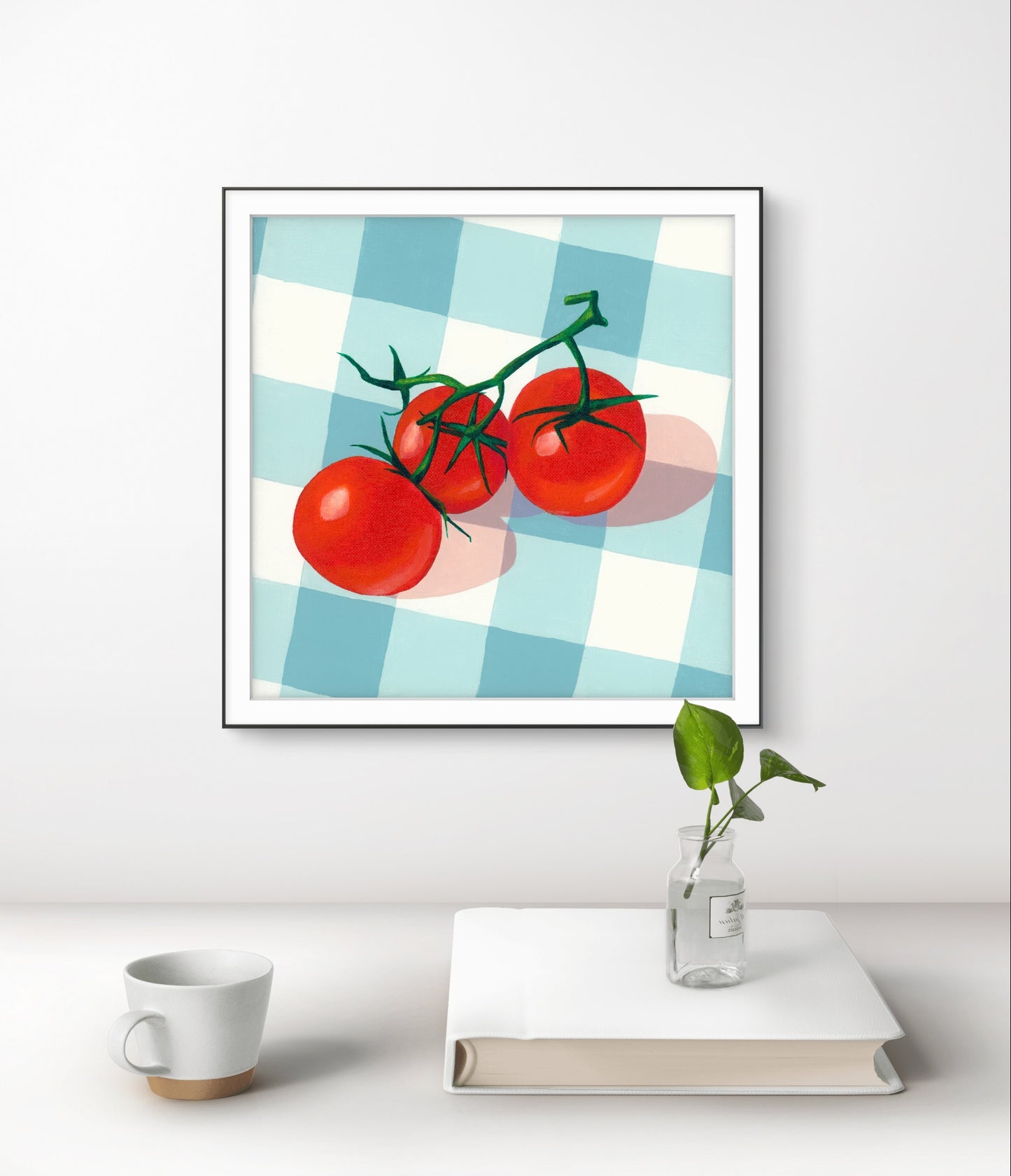 Framed artwork of tomatoes on a checkered background above a white surface with a cup, book, and plant.