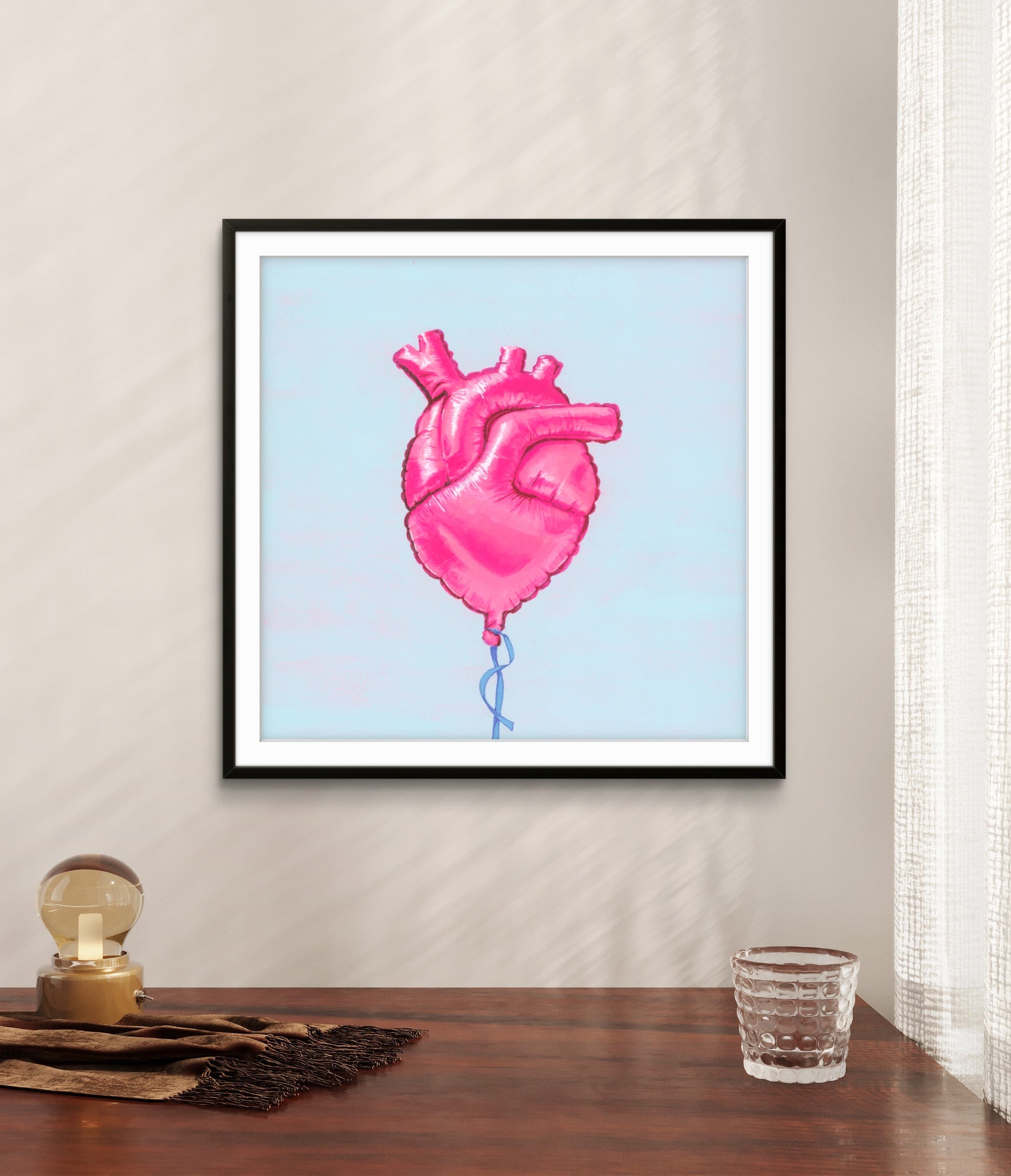 Framed artwork of a pink heart-shaped balloon on a light blue background, displayed on a wall above a wooden surface.