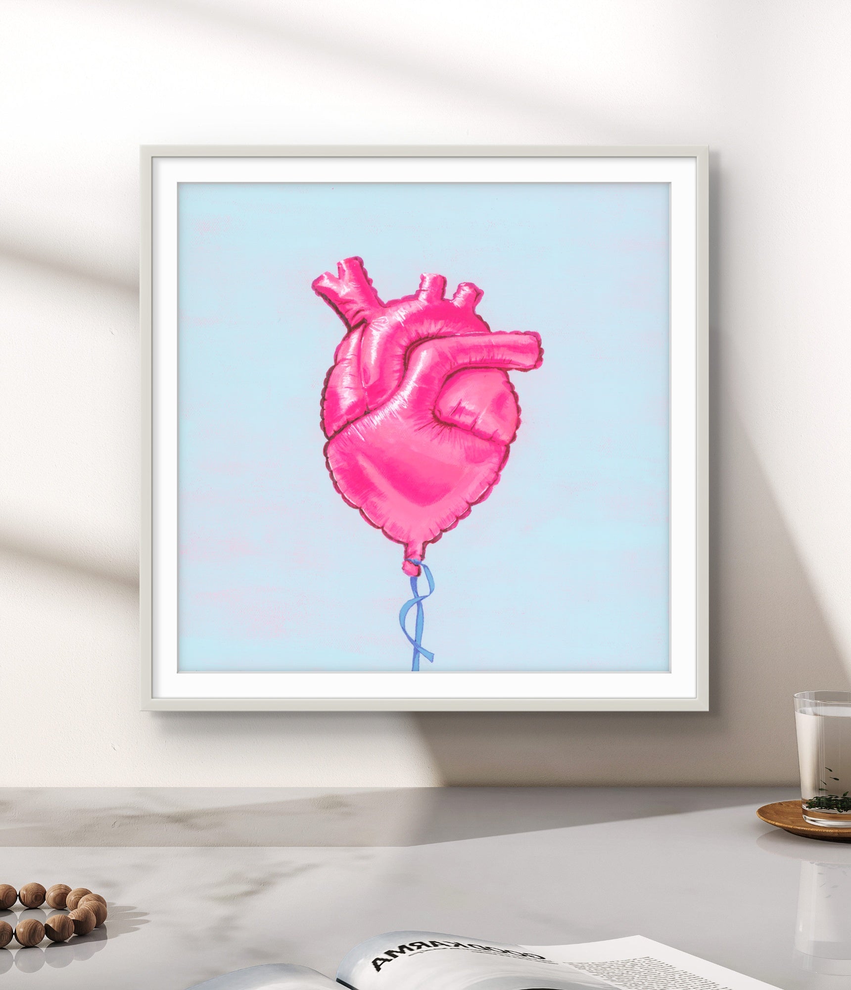 Framed artwork of a pink heart-shaped balloon on a light blue background, displayed on a wall.