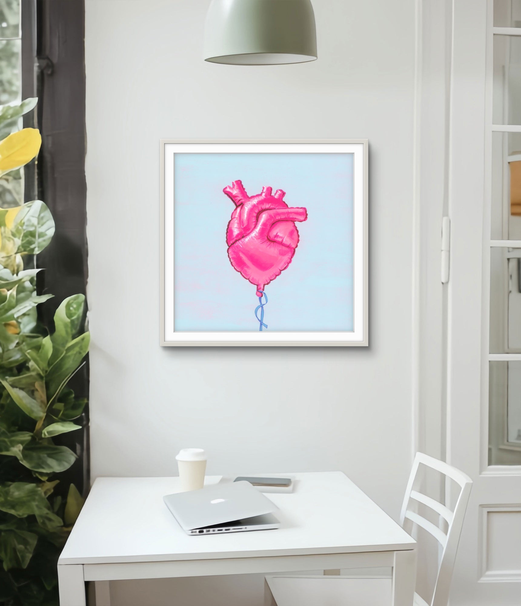Framed artwork of a pink heart balloon on a wall above a desk with a laptop and cup.
